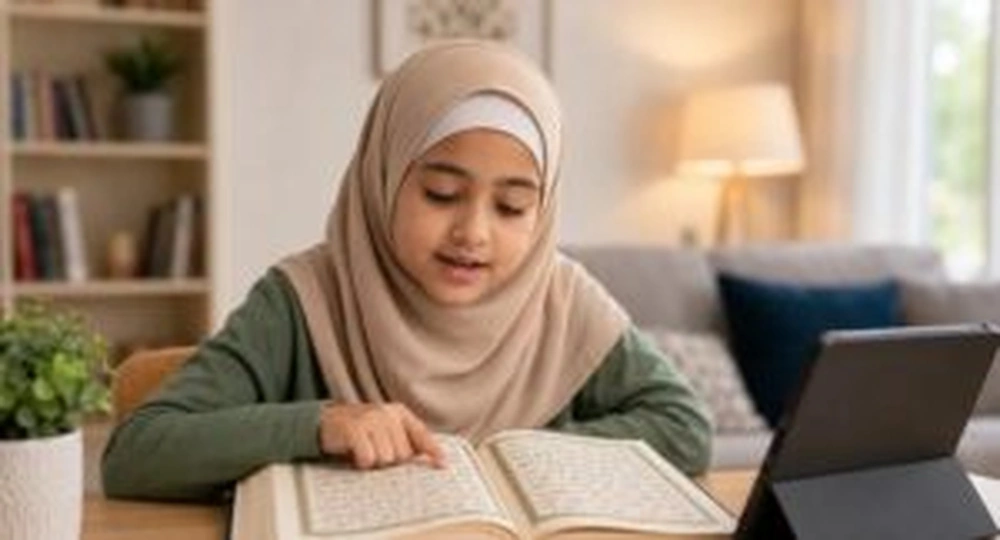 A woman wearing a hijab reading an open Quran beside a tablet at home